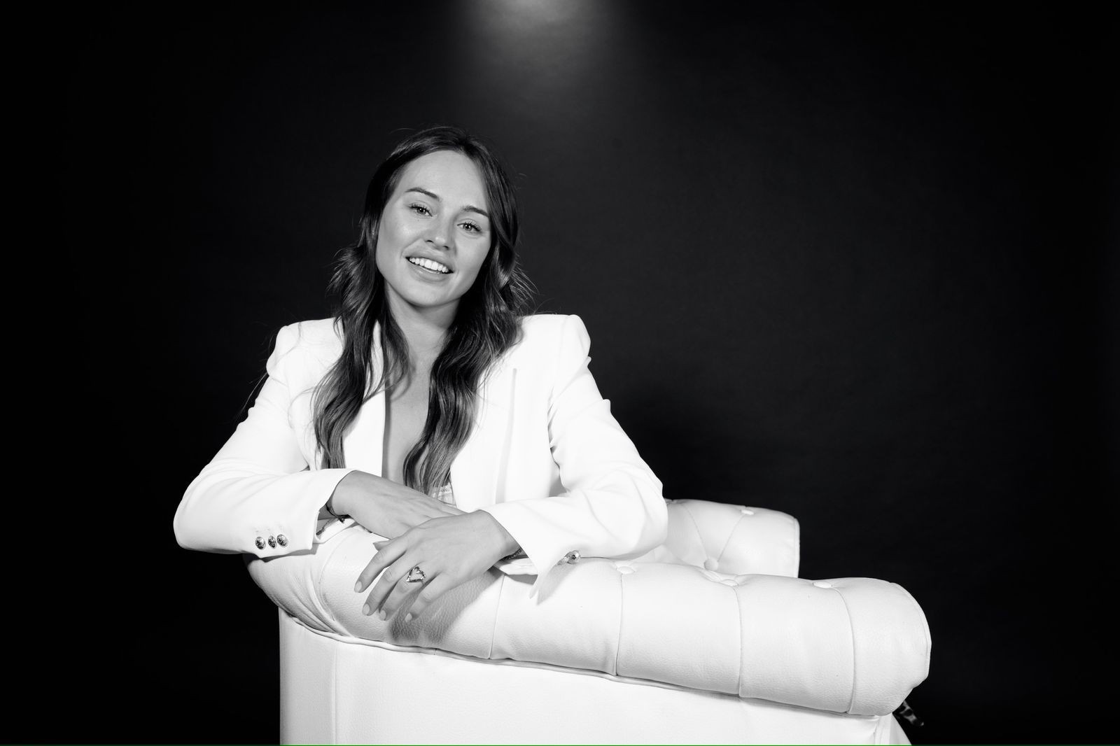 Person in a white suit seated on a white tufted chair against a dark background.